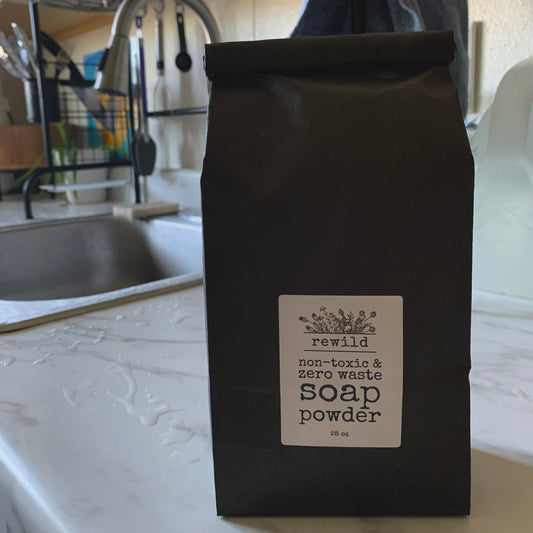 a black paper bag labeled "rewild non-toxic and zero waste soap powder 28 oz" sitting on a white marble kitchen counter with a sink in the background