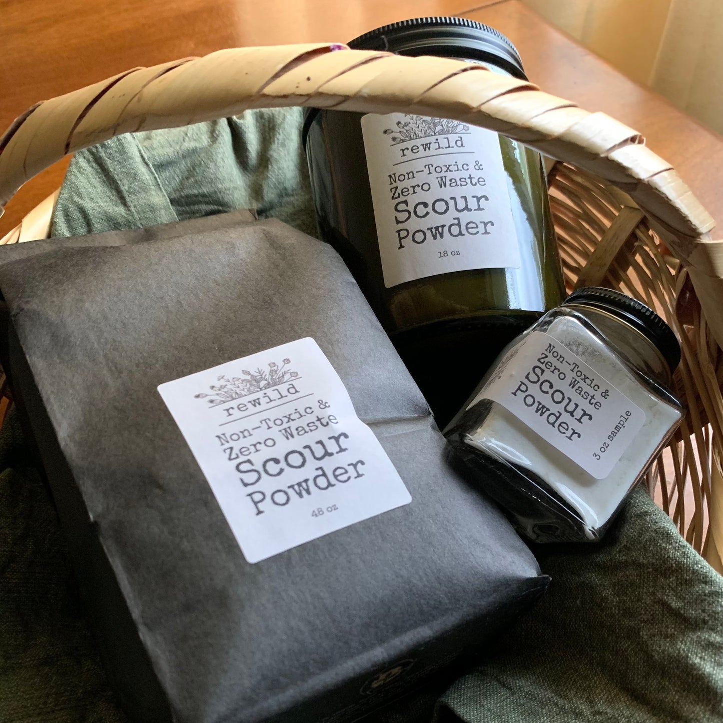 3 different sizes of scour powder (3 oz sample jar, 18 oz jar, 48 oz refill bag) in a woven basket