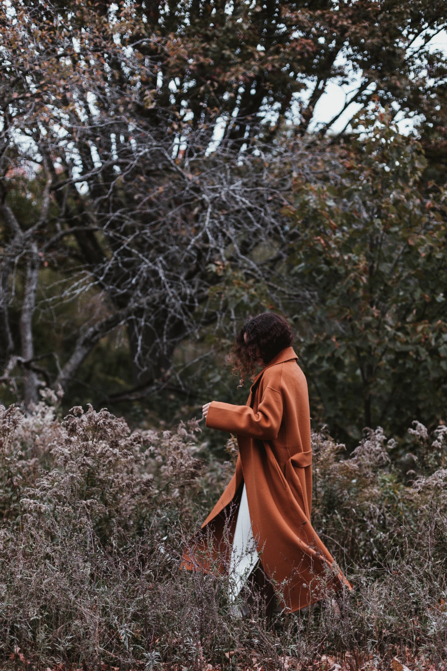 woman in long burnt orange coat in a woodsy field