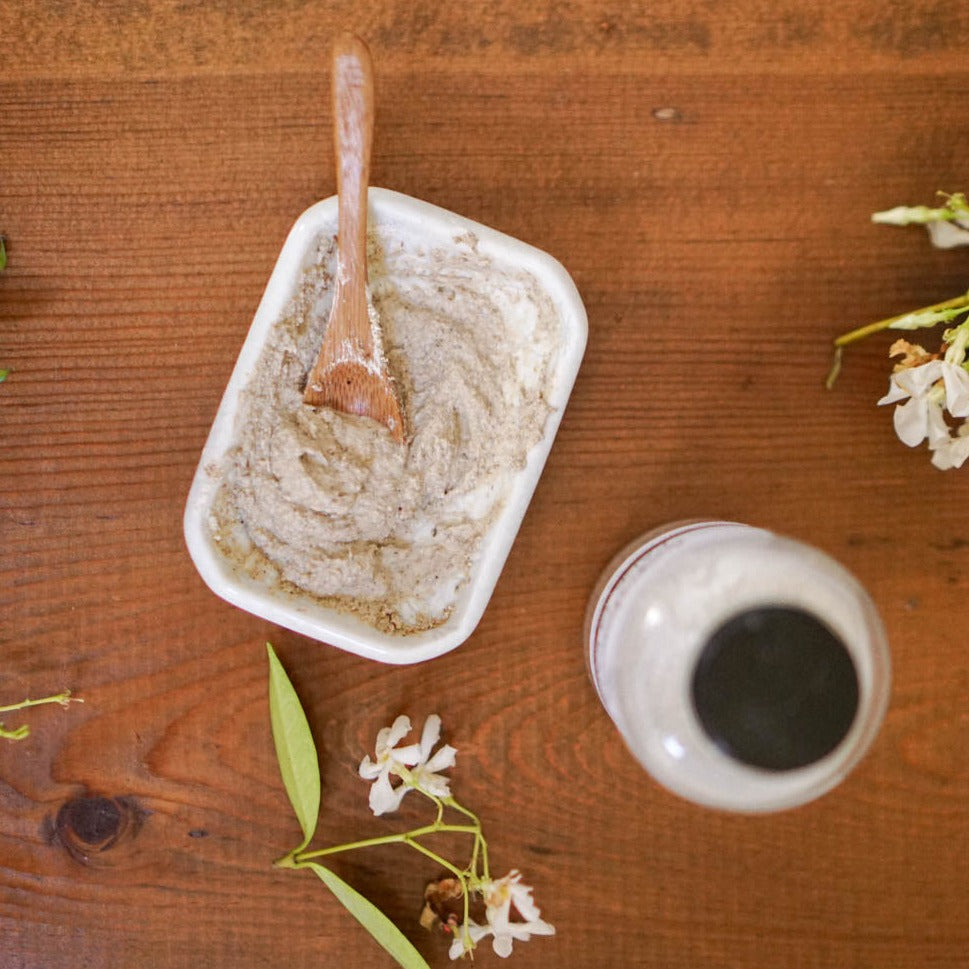 a rectangular ceramic dish of face mask powder ready to apply to face on a wood board with small flowers and a glass apothecary bottle