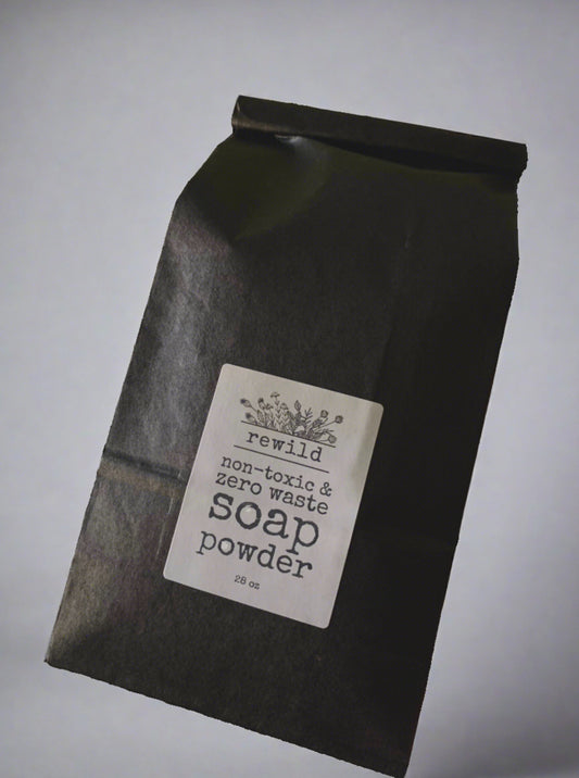 a black paper bag labeled "rewild non-toxic and zero waste soap powder 28 oz" on a white background