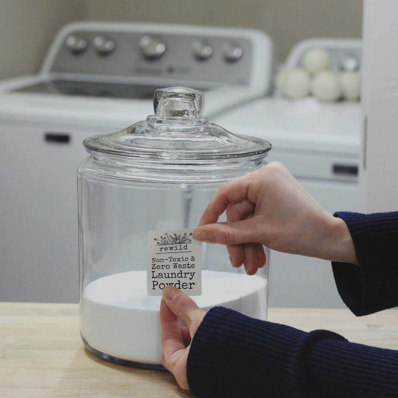 hands labeling a glass jar of laundry powder with washer and dryer in background