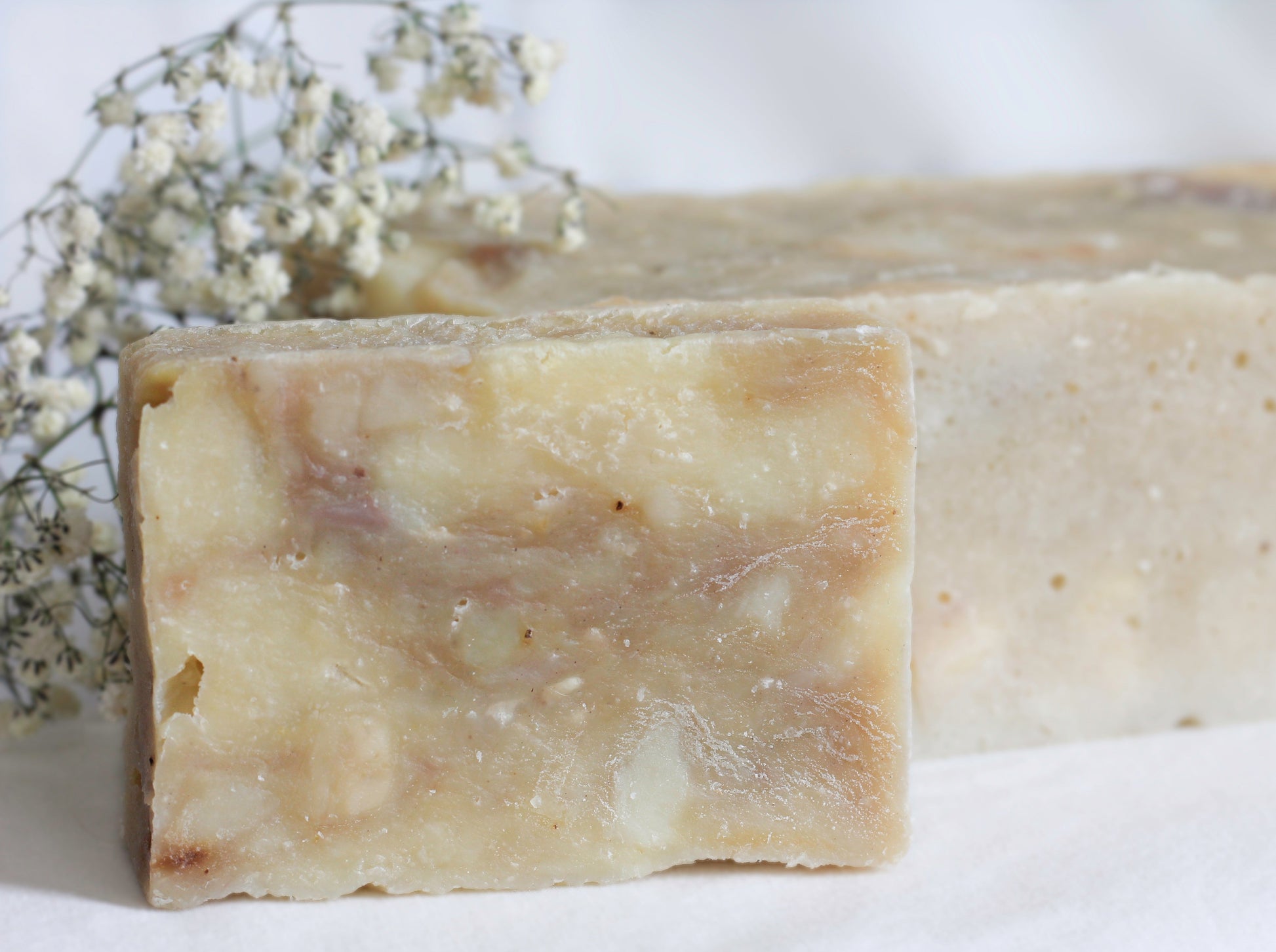 bar of soap in front of a loaf of soap and small white dried flowers on a white background