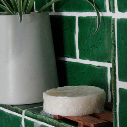 a lathered-up solid shampoo bar on a wood soap dish in a green-tiled shower