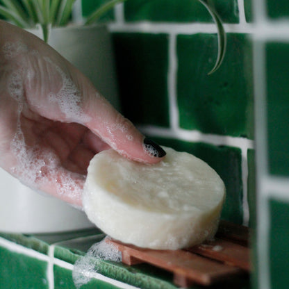 a hand placing a lathered-up solid shampoo bar on a wood soap dish in a green-tiled shower